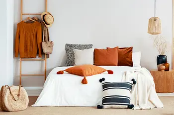 Modern eingerichtetes Schlafzimmer mit dekorativen Kissen in Erdtönen auf einem weißen Bett, kombiniert mit Naturmaterialien wie Basttasche, Holzleiter und Hängelampe.